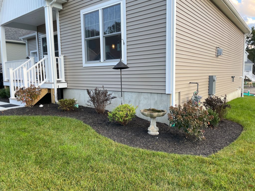 Corner garden bed with small flowering perennials, a young tree, and decorative birdhouse hanging on a branch near a white fence.